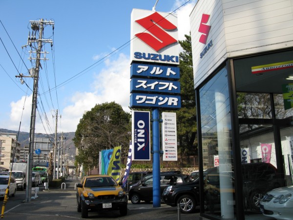 The Suzuki/used car dealer in Kobe. Under the Suzuki sign, many dealers list the cars they sell. Here, it states the Alto, Swift, and Wagon R.